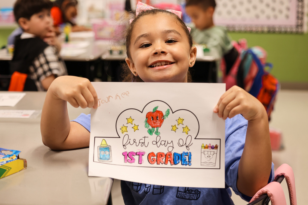 A smiling girl holds a white sign with colorful images, text, and illustrations. She is seated at a desk with other students.
