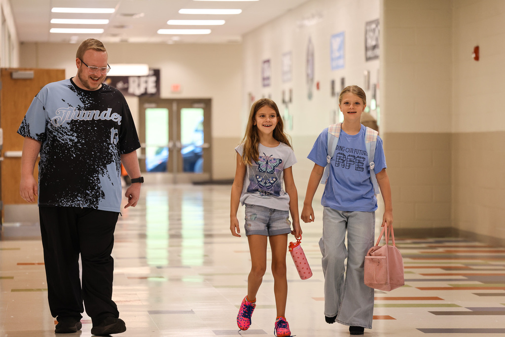 An adult and two children walk through a school hallway. The adult wears glasses and a t-shirt.