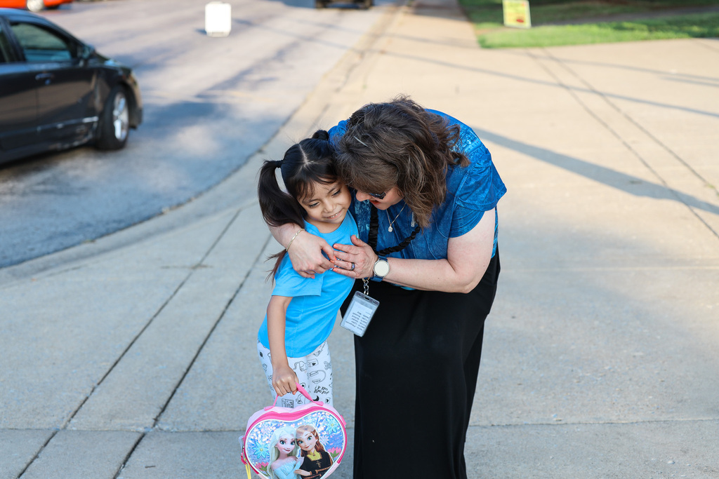 Woman in blue top hugs girl with ponytail, carrying pink backpack with Frozen characters, on sidewalk.