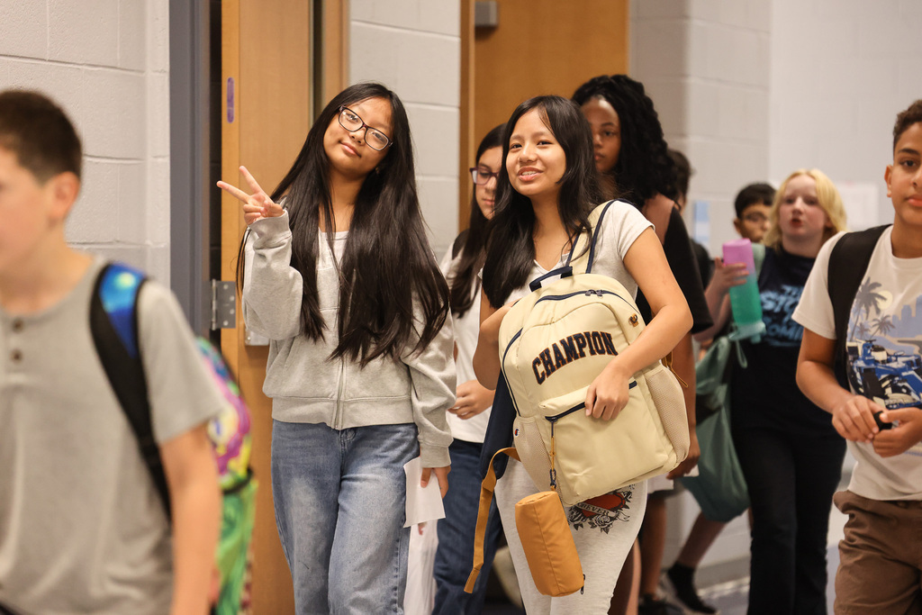 Students in a hallway with backpacks; one girl gives a peace sign, another carries a "CHAMPION" backpack.