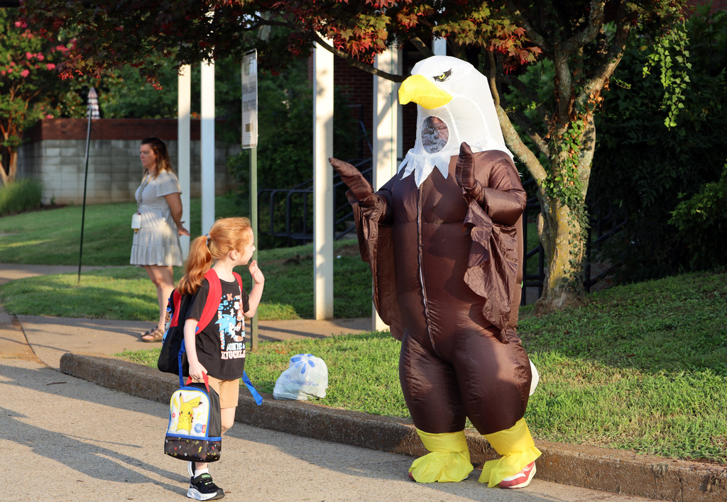 A person in an eagle costume waves to a child carrying a backpack on a sidewalk.