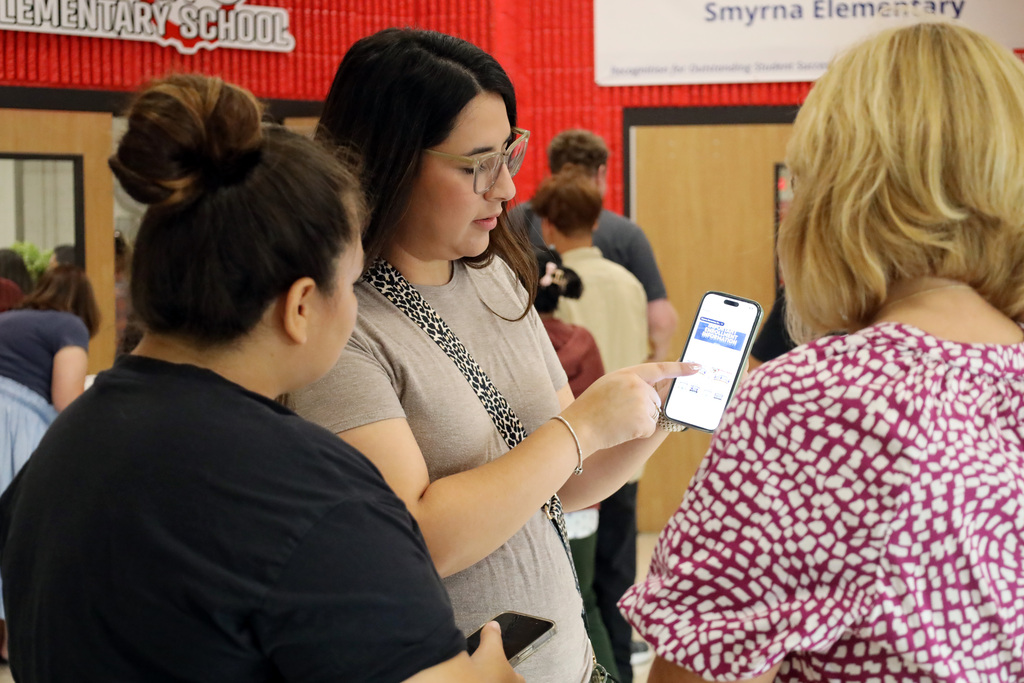 Three women with phones, one holding a phone, inside a room. Behind them, a wall with text.