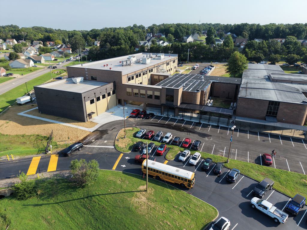 Aerial view of a school with a yellow school bus in the parking lot. Buildings, cars, and trees in the background.