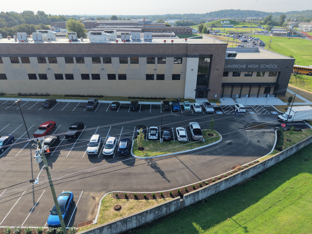 Aerial view of a school building with a parking lot, vehicles, and a grassy field.