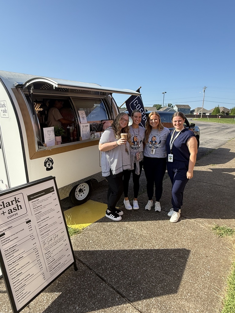 Four people stand beside a food truck, smiling and holding cups. A menu board is nearby. A car and trees are in the background.