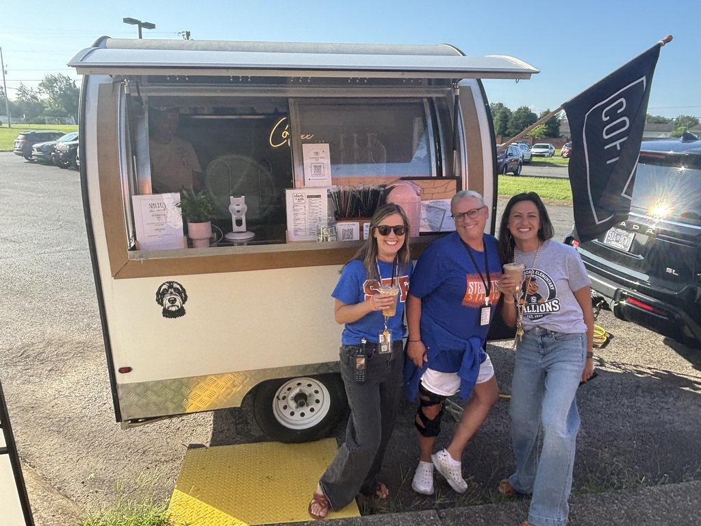 Three people pose in front of a food truck. One holds a drink and others wear blue shirts.