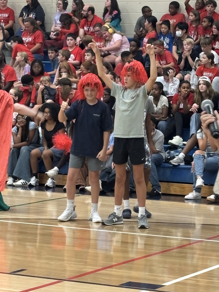 Two young people with red wigs stand on a basketball court, raising their hands. A crowd sits in the stands.