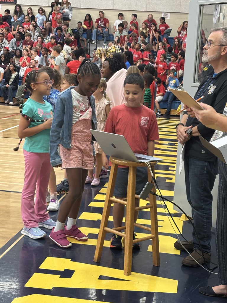 Children stand around a laptop on a wooden stool on a basketball court. A man in glasses holds papers.