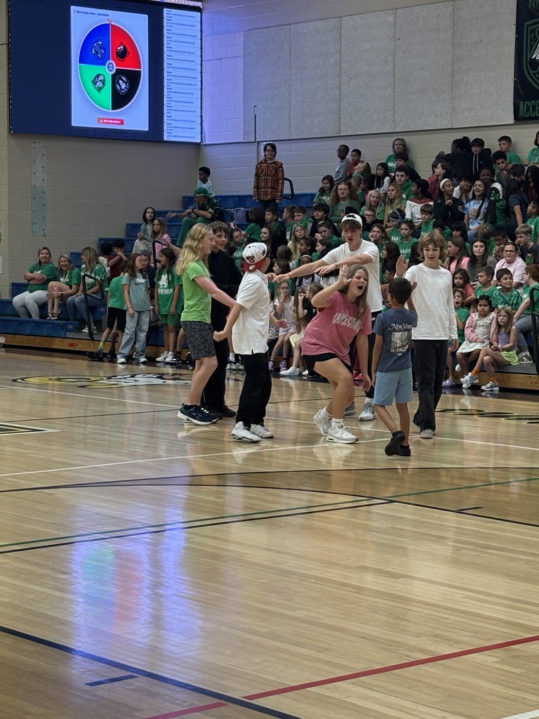 A group of people on a wooden basketball court, some standing and others sitting in bleachers.