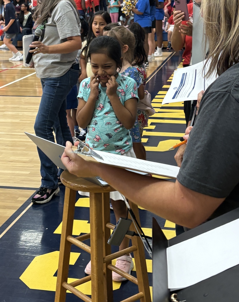 A young girl stands on a stool as an adult holds a clipboard and paper. The background shows a gymnasium.