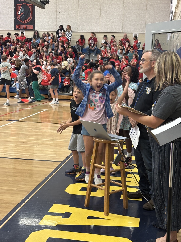 A group of people stands on a raised platform inside a gym. A girl stands on a stool with a laptop, raising her arms.