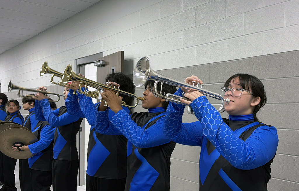 A group of musicians in blue and black uniforms play trumpets in a room with a white wall.