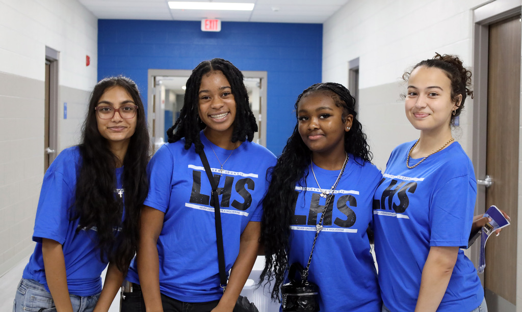 Four young women in matching blue shirts pose in a school hallway. They wear necklaces and bags.