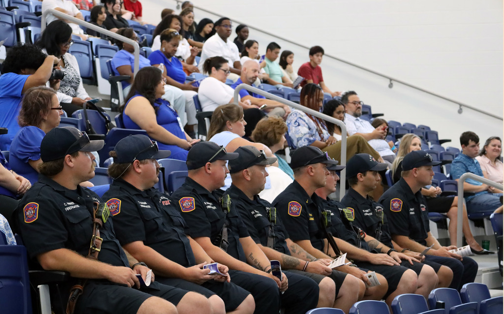 Eight firefighters in uniform sit in blue chairs at an event. They wear caps and some have name tags.
