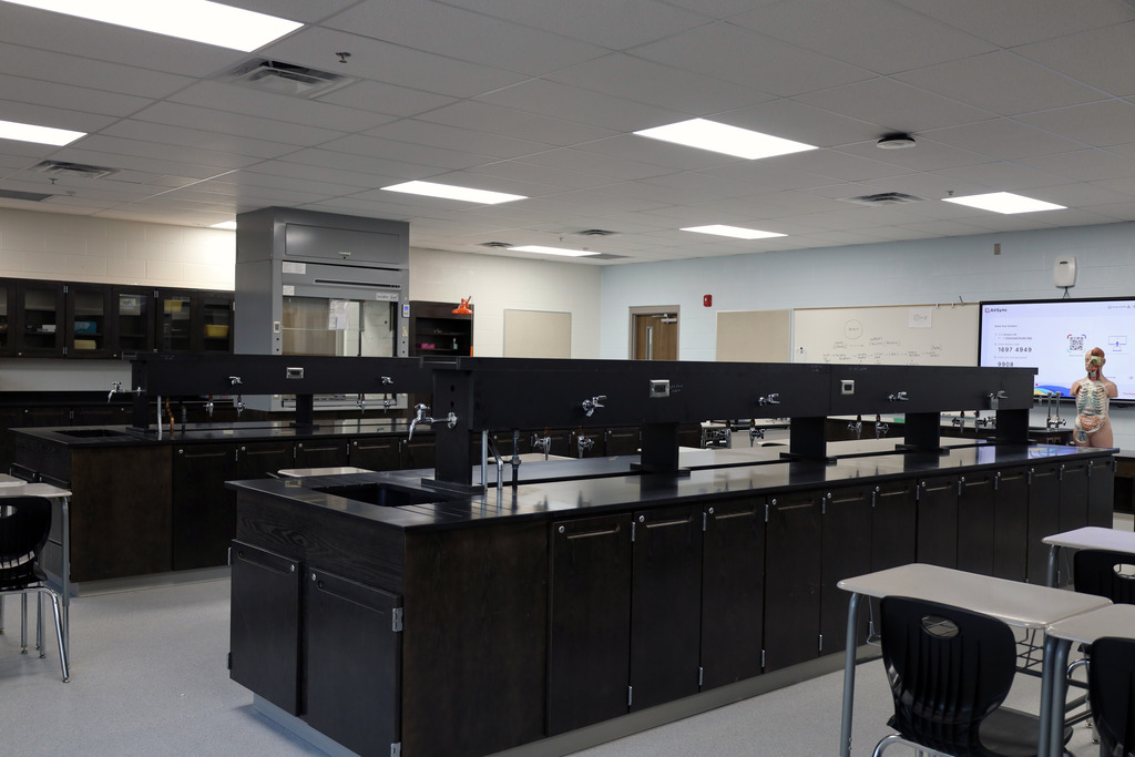 A laboratory with long counters, sinks, and cabinets. Overhead lights and a screen on the wall. A person stands near a white board.