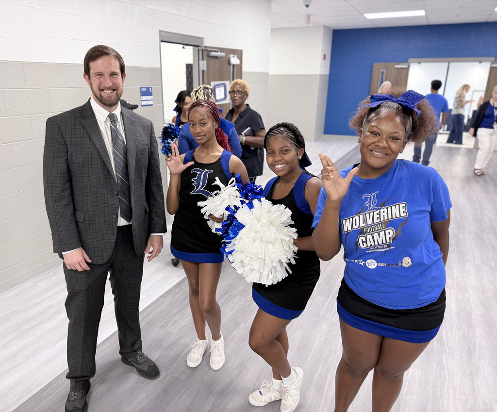 Man in business attire stands with three women in cheerleading uniforms in a school hallway.