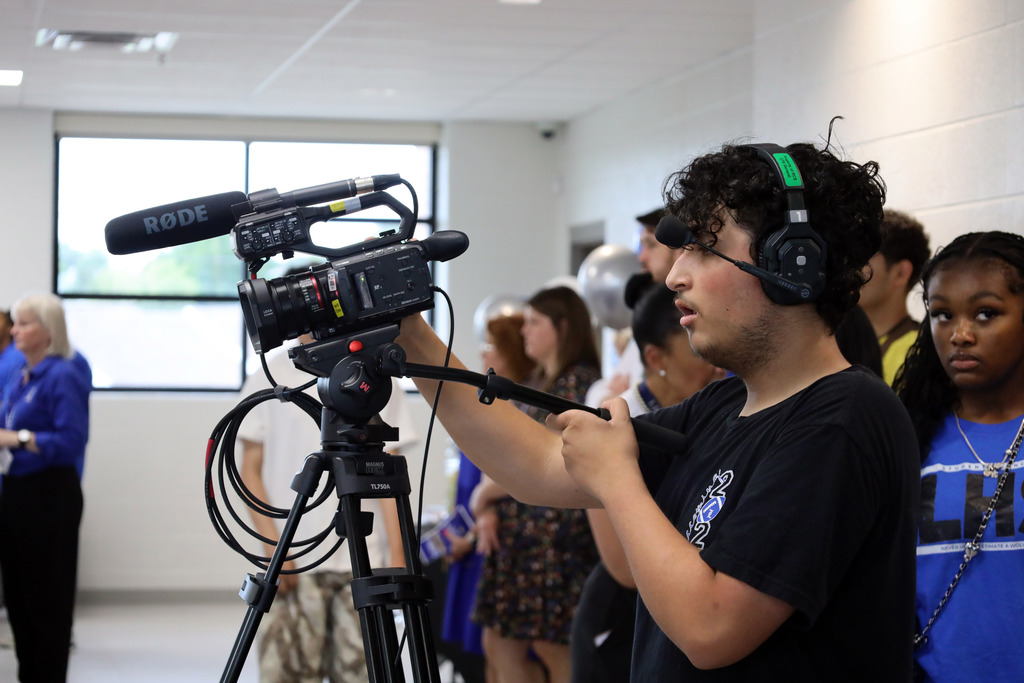 A person in a black shirt holds a video camera while several others stand in the background.
