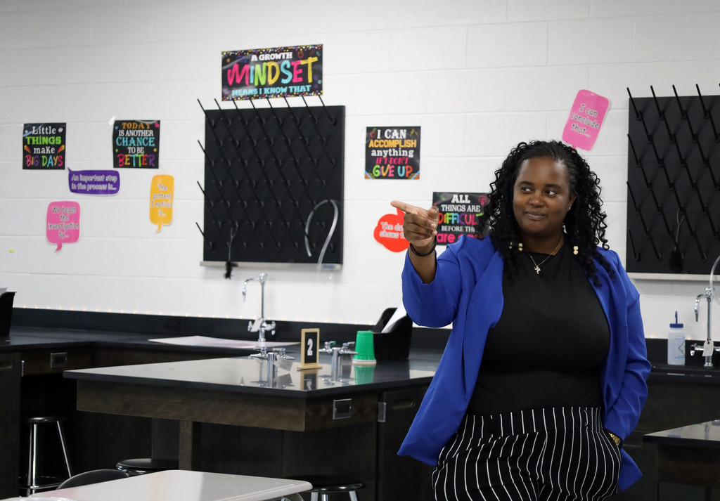 A woman with curly hair points to the wall in a classroom with motivational posters.