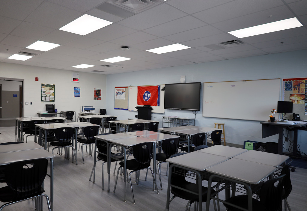 An empty classroom with desks and chairs arranged. A whiteboard and a large screen are on the wall.