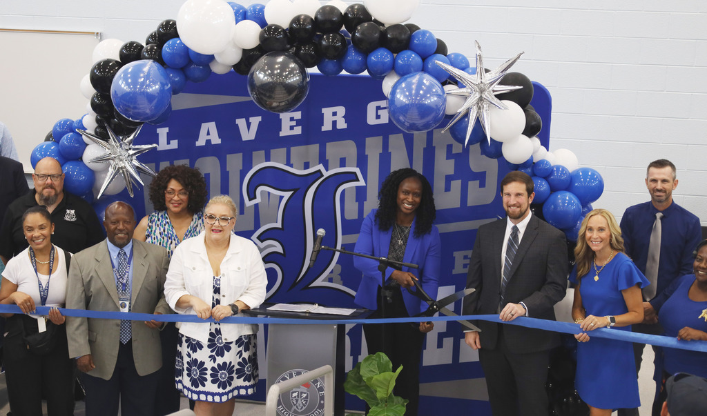 A group of adults poses behind a blue and white backdrop with balloons and a ribbon.