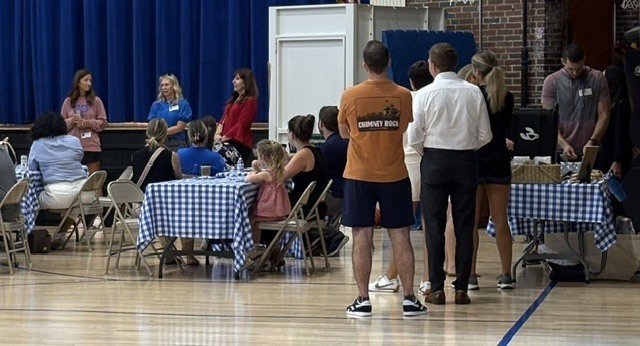 People in a room with a blue curtain. Some sit at tables with blue-and-white cloths, others stand.