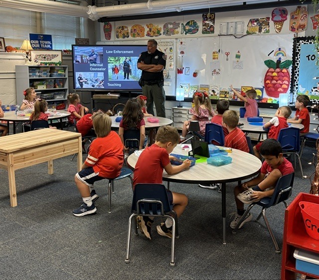 A classroom with children in red shirts seated at tables. An adult stands at the front by a screen.