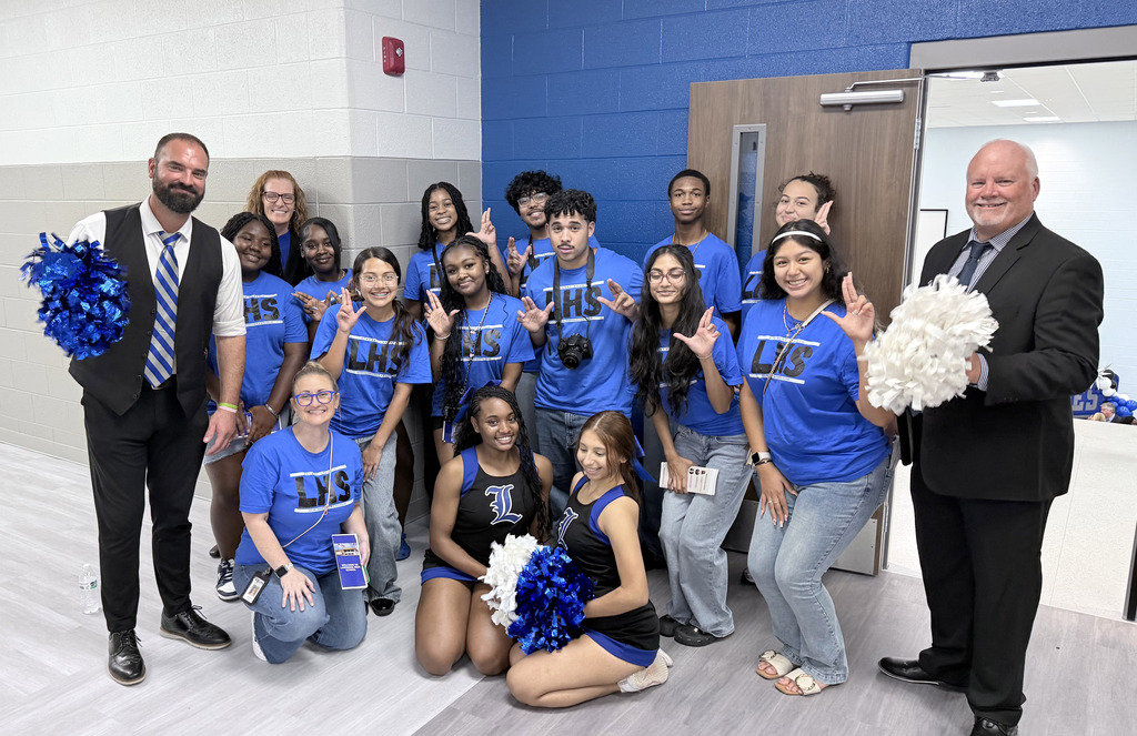 Cheerleaders in blue uniforms and two men, one with a blue pom-pom, pose in a blue-walled room.