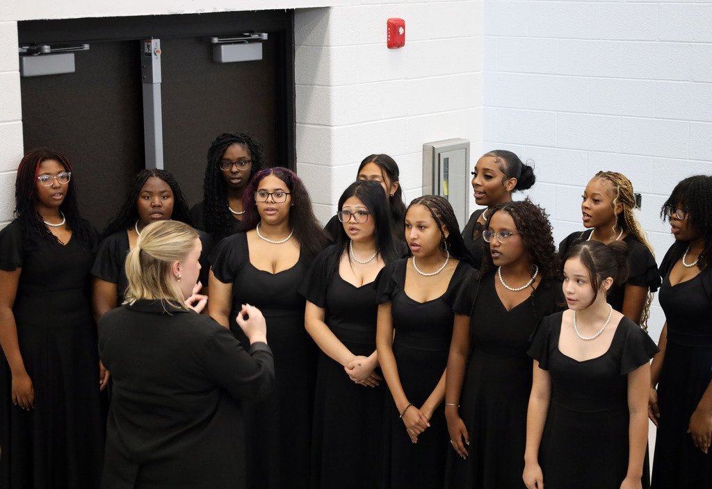 A group of women dressed in black, some wearing necklaces, standing in a row, with a woman in front of them.