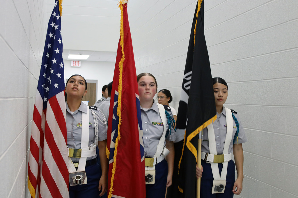 Four people in matching uniforms stand in a hallway with white walls, holding flags. A woman looks down.