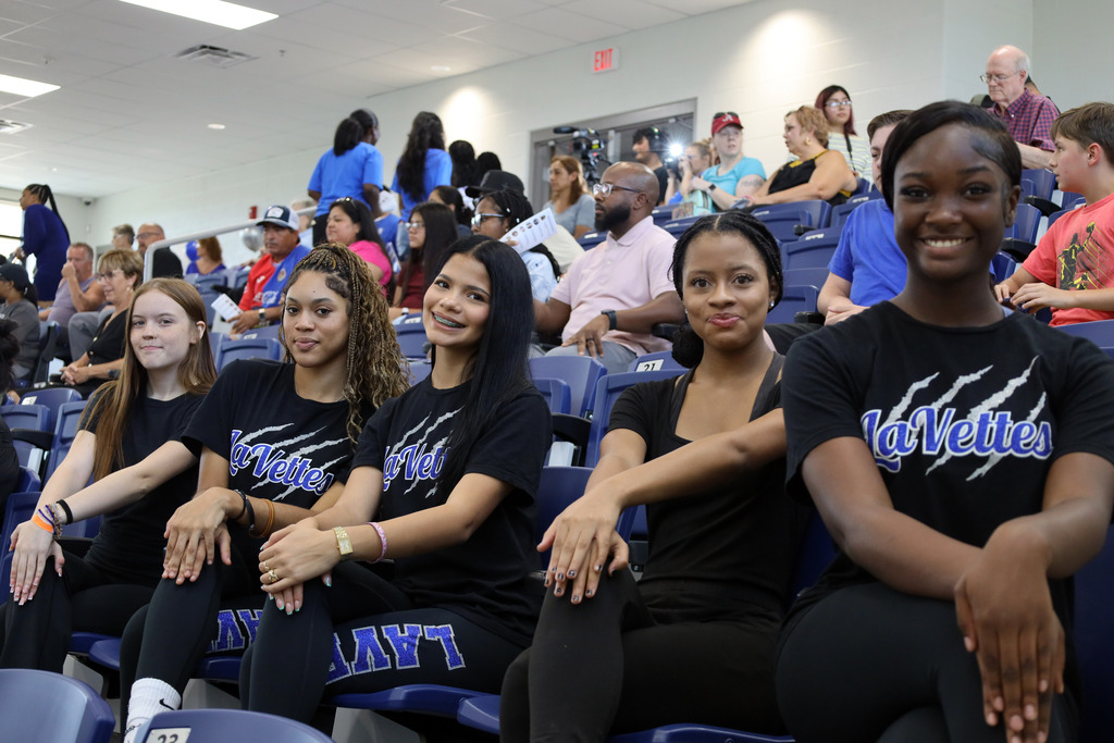 A group of five women wearing black shirts sit in the audience of a stadium. They are smiling and looking in various directions.