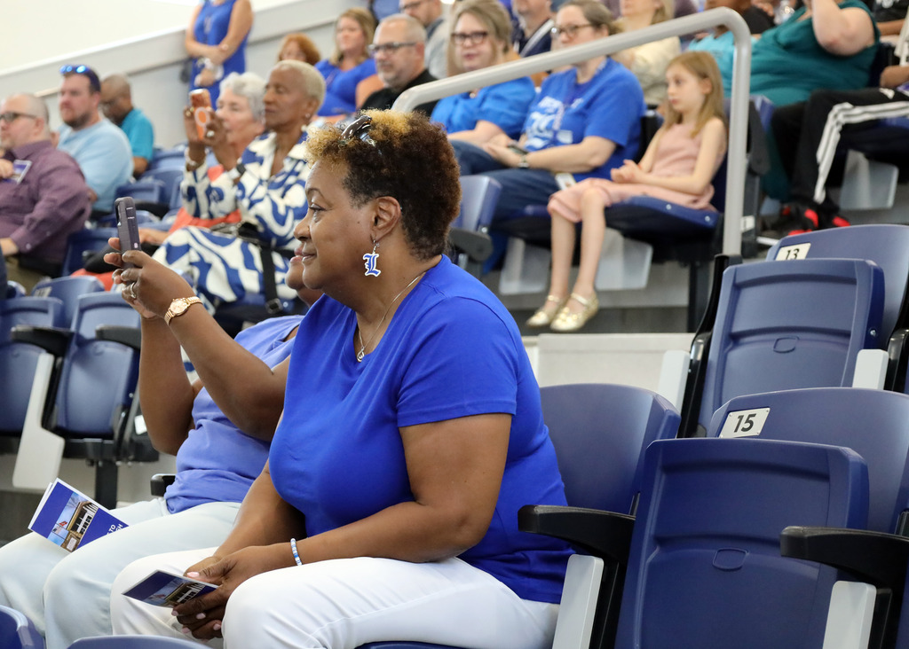 People seated in a stadium with blue chairs, one woman capturing a moment with a phone.