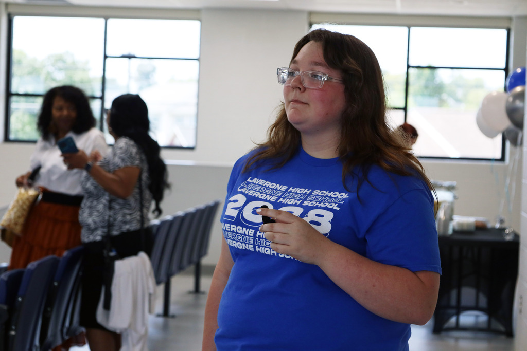 A person in a blue shirt labeled "2028" holds a phone indoors with others, rows of chairs, and windows.