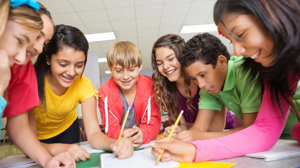 A group of children are gathered around a table, looking at papers and writing with pencils.
