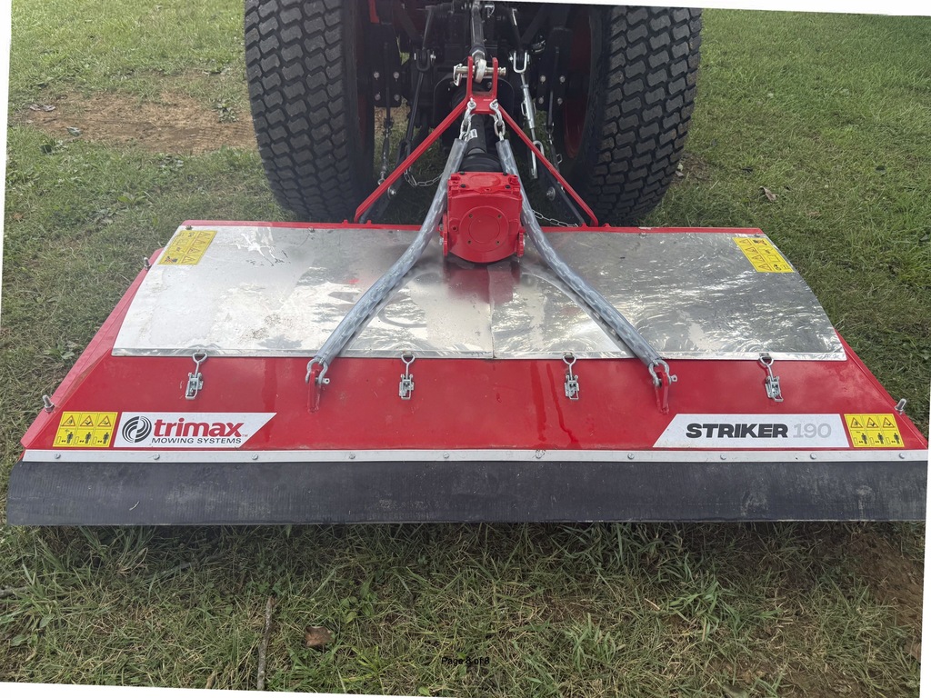 A red and gray lawn mower attachment labeled "TRIMAX" and "STRIKER" is on a grassy field.