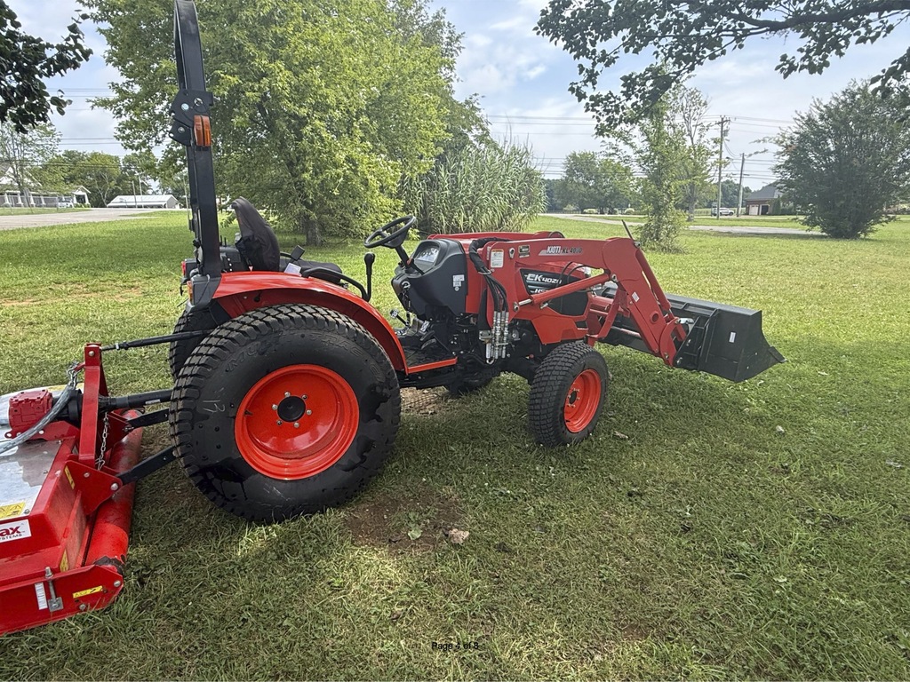 A red tractor with a black seat is parked on a grassy field. A red attachment is on the left. Behind the tractor are trees and a clear sky.