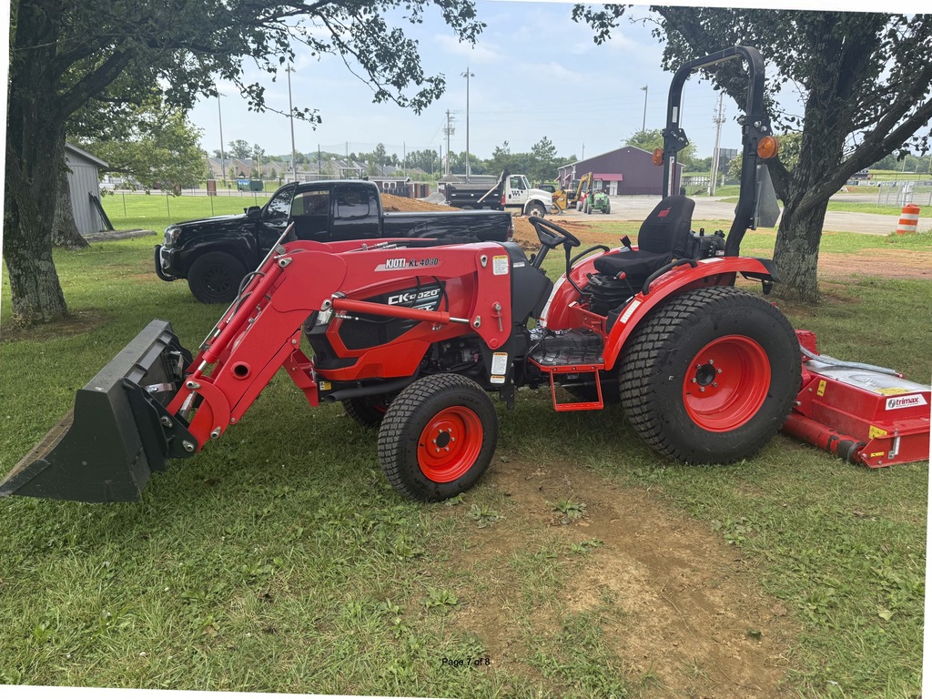 Red tractor parked in a field with trees and a black pickup truck in the background.
