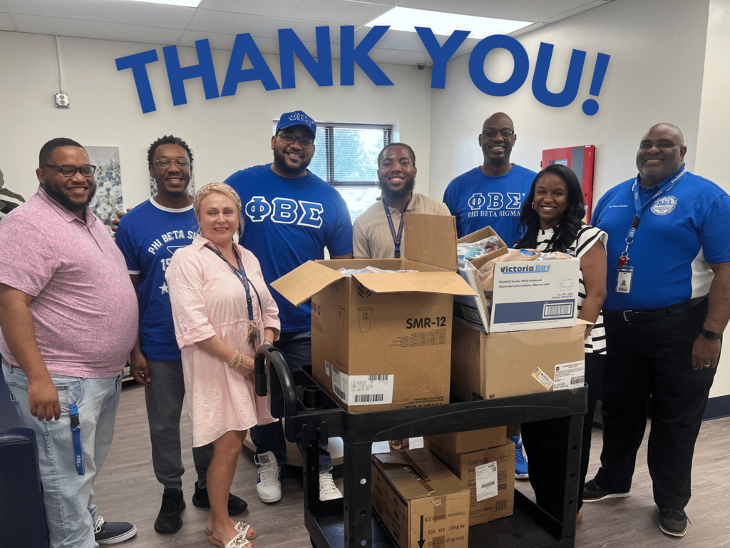 Group of people, both men and women, wearing blue shirts stand in a room with boxes, smiling.