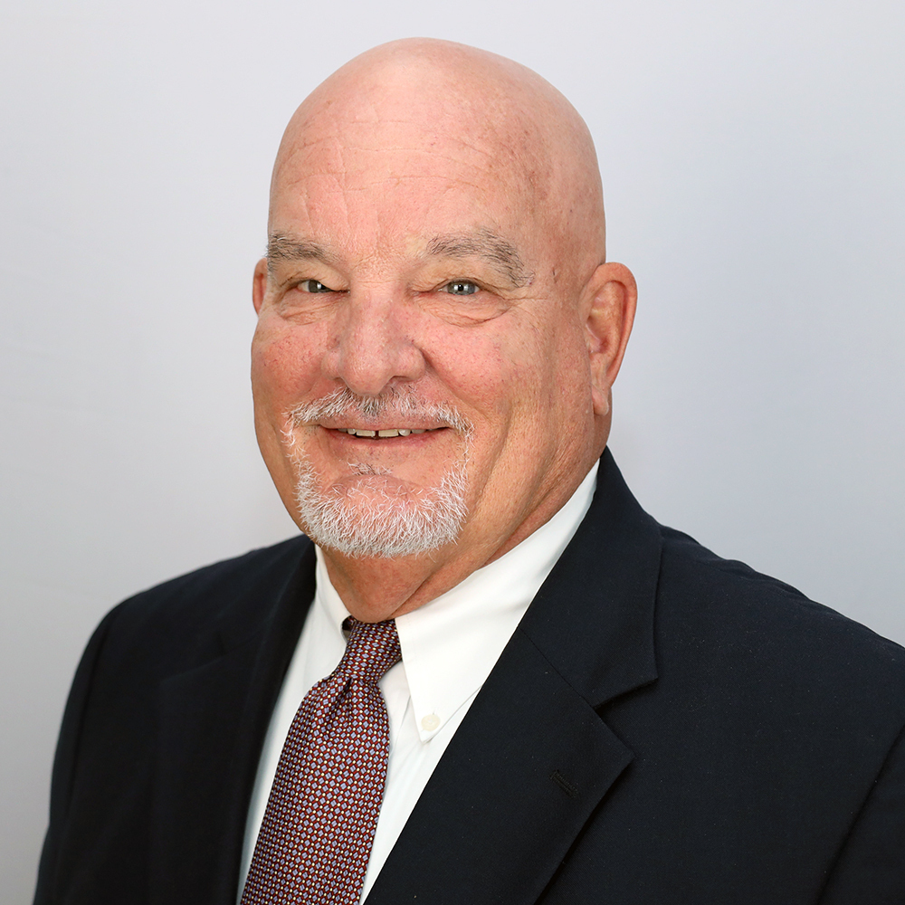 Man with a beard and mustache, smiling, wearing a black suit jacket and white shirt, and a brown tie.