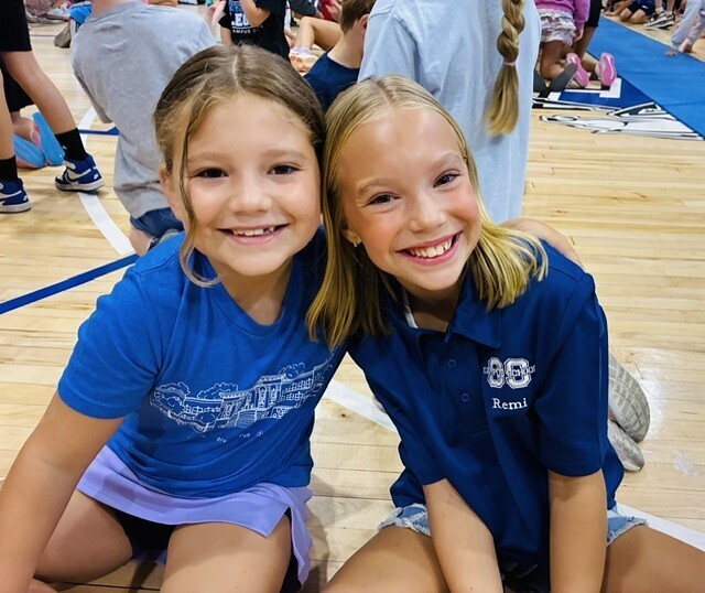 Two girls, both wearing blue shirts, sit on the floor in a gym. They are smiling and posing for a photo.
