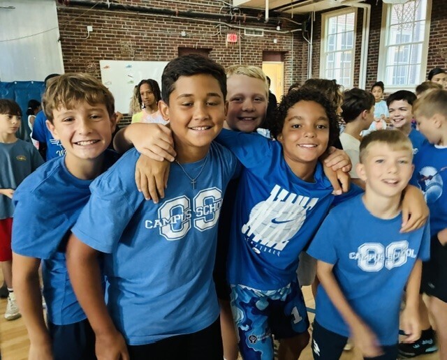 Group of kids wearing blue shirts with "CAPES" and numbers. Some have hands on shoulders. Background shows brick wall, glass windows, and people.