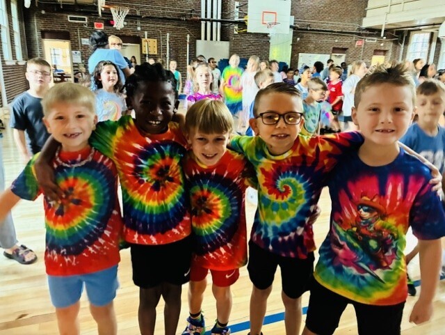 Five children in tie-dye shirts pose for a photo in a gymnasium. Several other children are in the background.