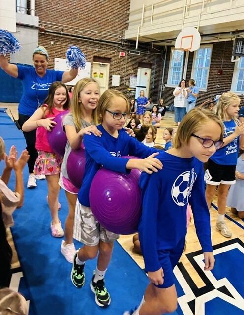 Children holding purple balloons in a gymnasium. A woman in a blue shirt stands behind them.