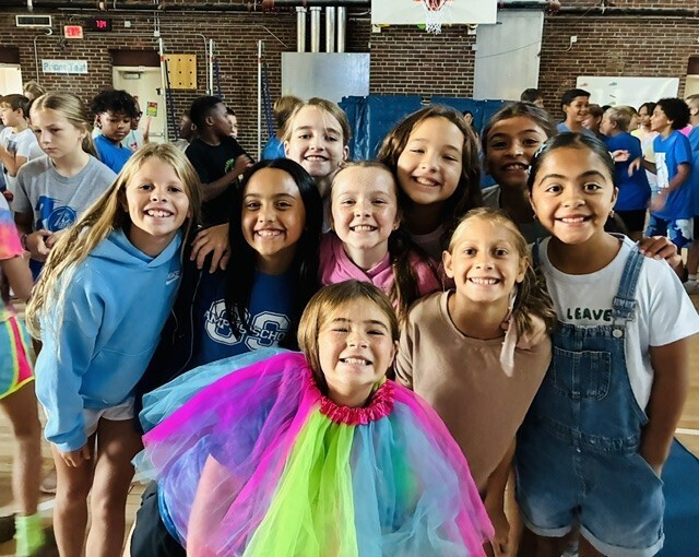 Group of children in a gymnasium, smiling for a photo. One wears a colorful tutu.