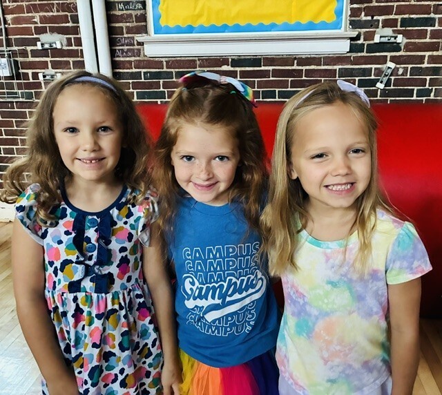 Three girls stand together, smiling, with one girl wearing a blue shirt with text. A red bench and brick wall in the background.