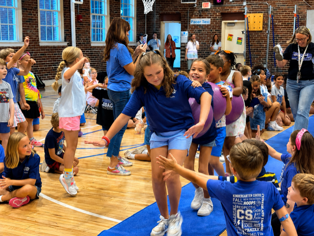 A group of children in a gymnasium, wearing matching blue shirts, play on a blue mat.