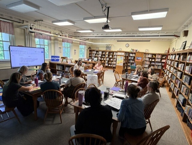 Group seated around tables in a library. A projector screen displays information in the background. Books are on shelves.