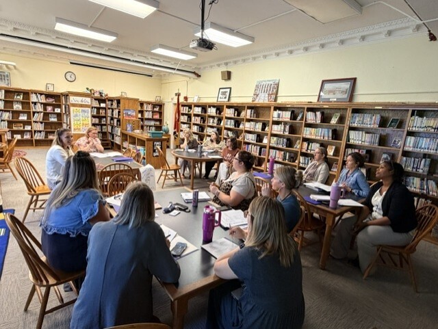 A group of women in a library. Some sit at tables, others stand, all engaged in conversation.