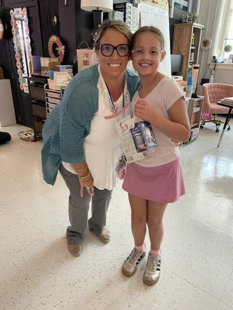 A woman and a girl in glasses and a pink skirt hold items and stand in a classroom.