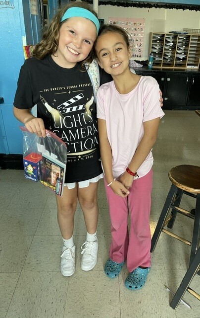 Two girls pose in a room with a blue door and brown stool. One holds a bag and the other wears pink pants.