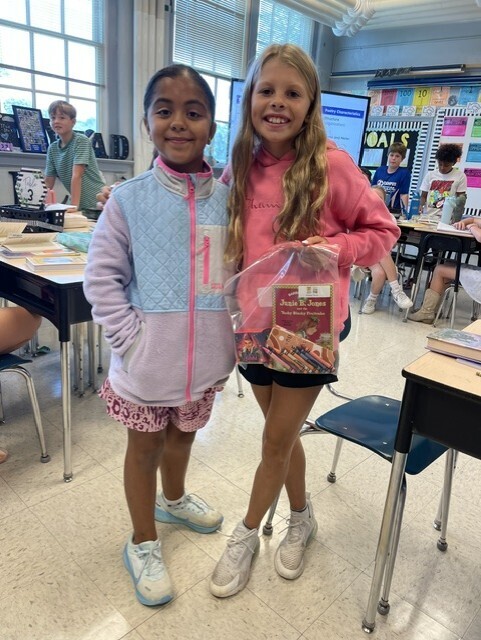 Two girls in jackets stand in a classroom, one holding a bag. Behind them, students sit at desks.